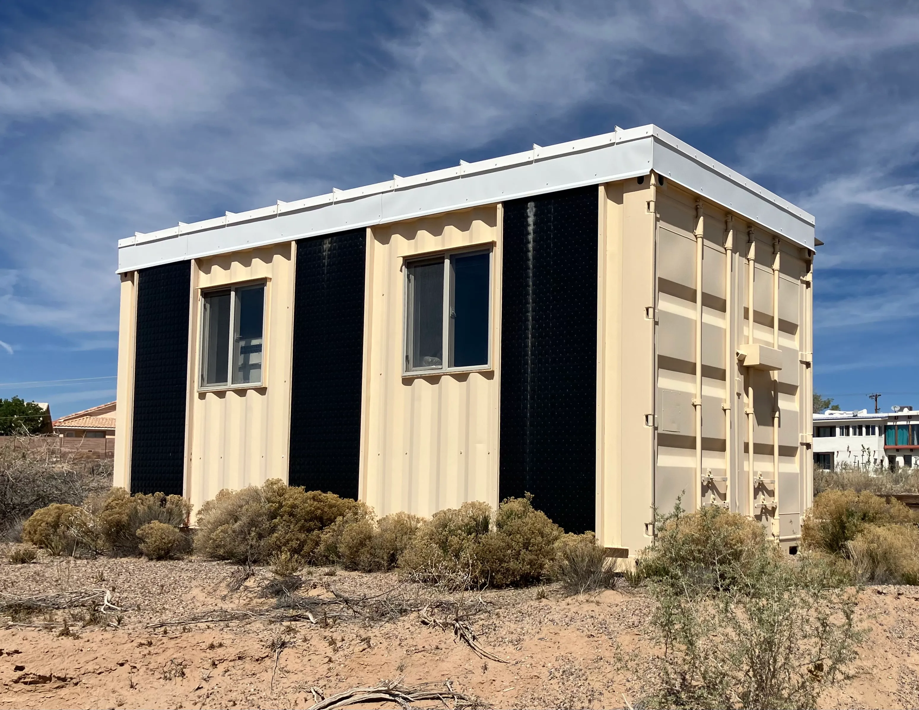 Exterior of a tan-painted shipping-container building with vertical black thermal-absorber panels between two windows, in a high-desert lot