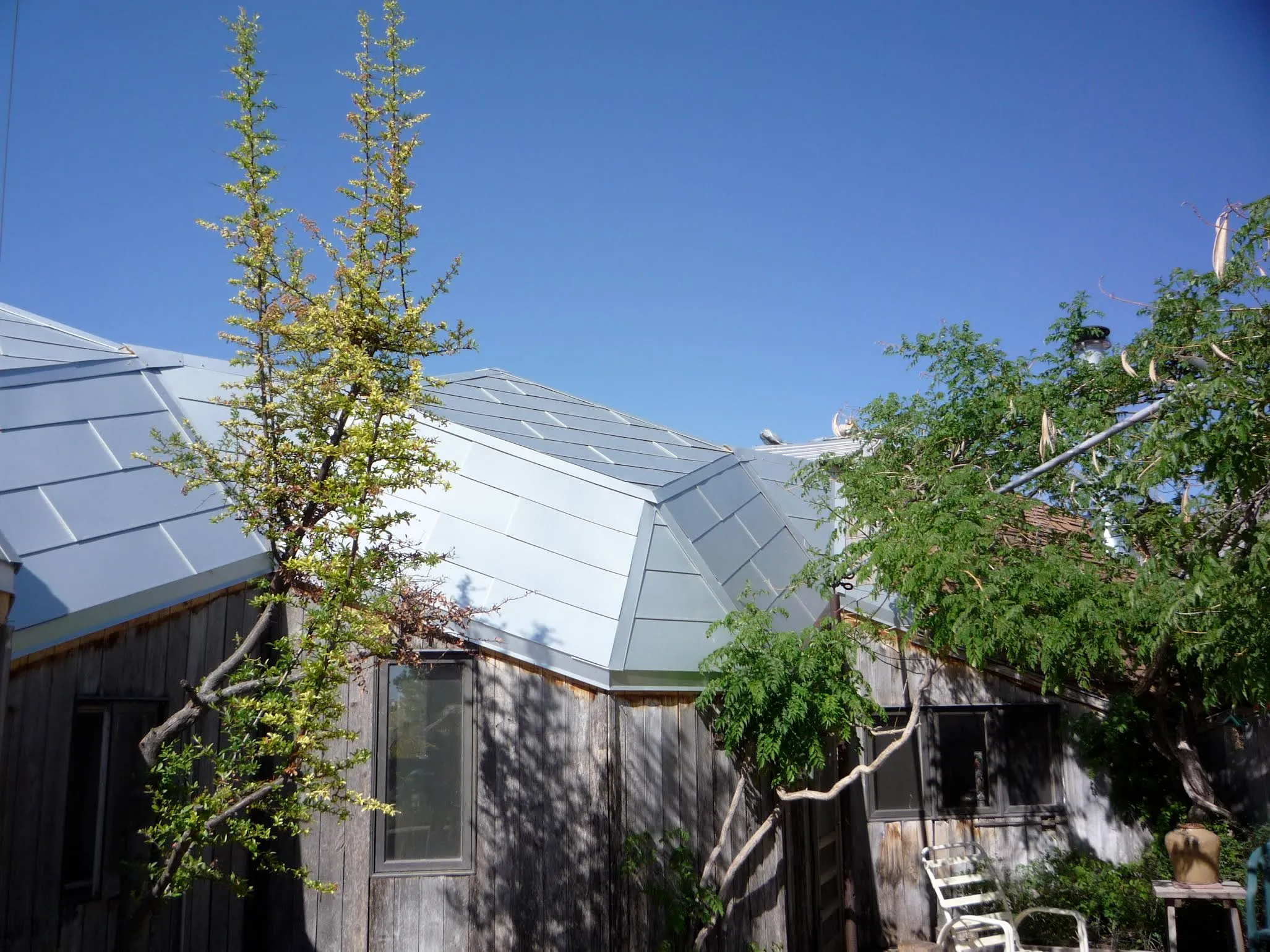 North view of the zome structure showing weathered wood siding and triangular aluminum roofline with desert trees in foreground
