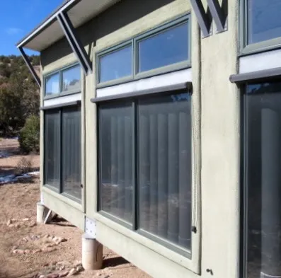 South-facing exterior of the Sol-Mod modular house: sage-green stucco wall with clerestory awning windows under a low roof, tall fixed glazing below revealing vertical translucent water-storage tubes, the box lifted on concrete piers in a snow-dusted piñon-juniper landscape