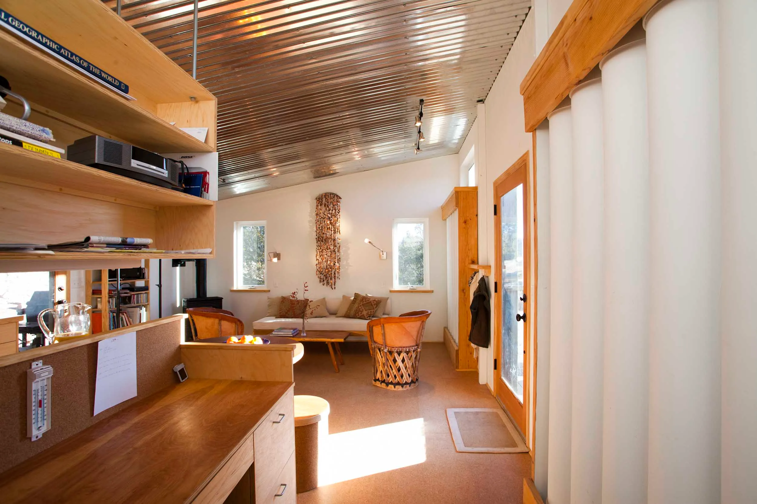 Interior of the Sol-Mod house photographed by Marc Romanelli: a narrow daylit room with a corrugated aluminum ceiling, vertical white water-storage columns along the right wall beside a glazed wood door, built-in birch desk and shelving on the left with a cork bulletin board and thermometer, cork flooring, leather equipal chairs and a sofa