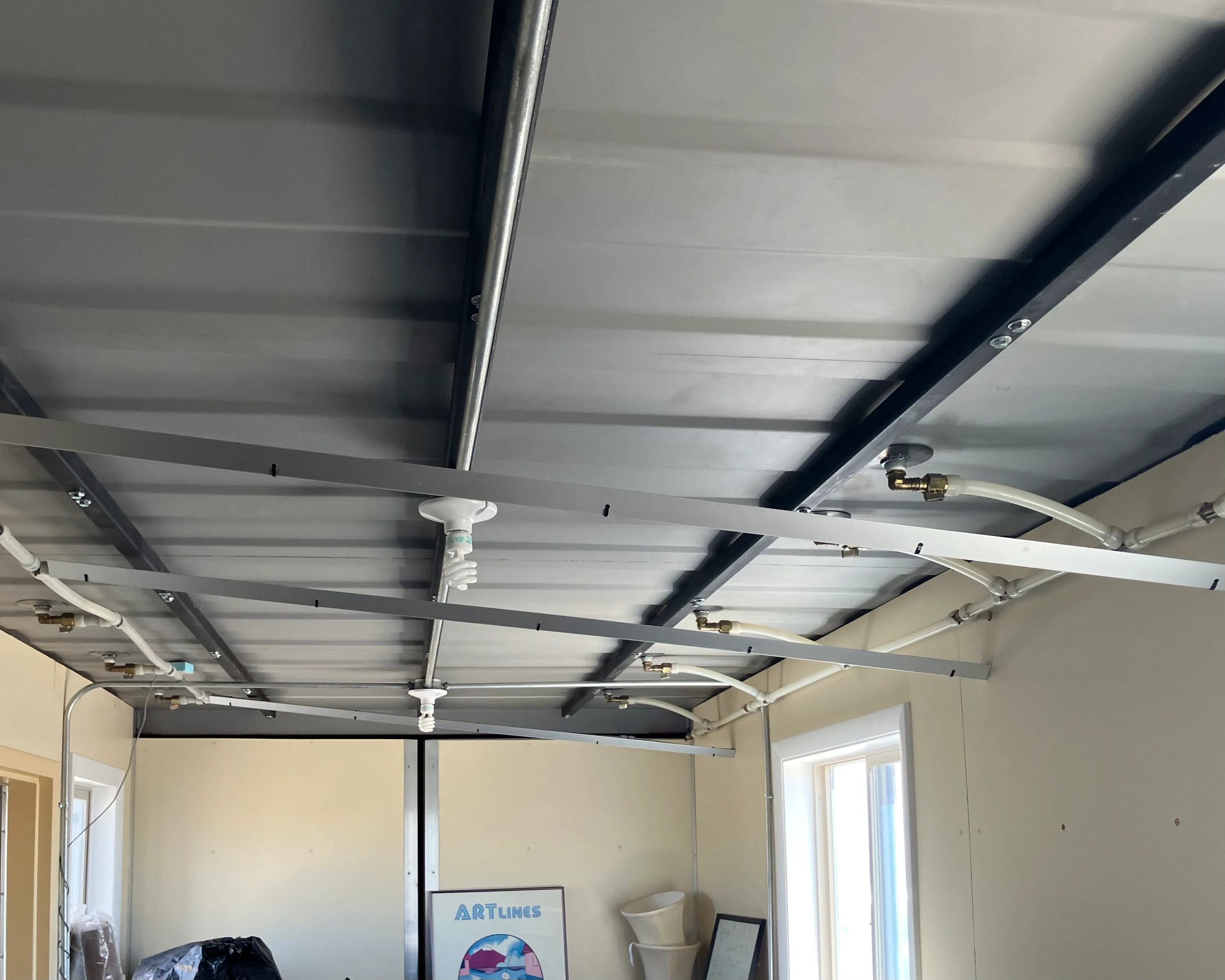 Interior view looking up at Double Play metal ceiling panels with hydronic piping, brass fittings, and a CFL lamp pendant