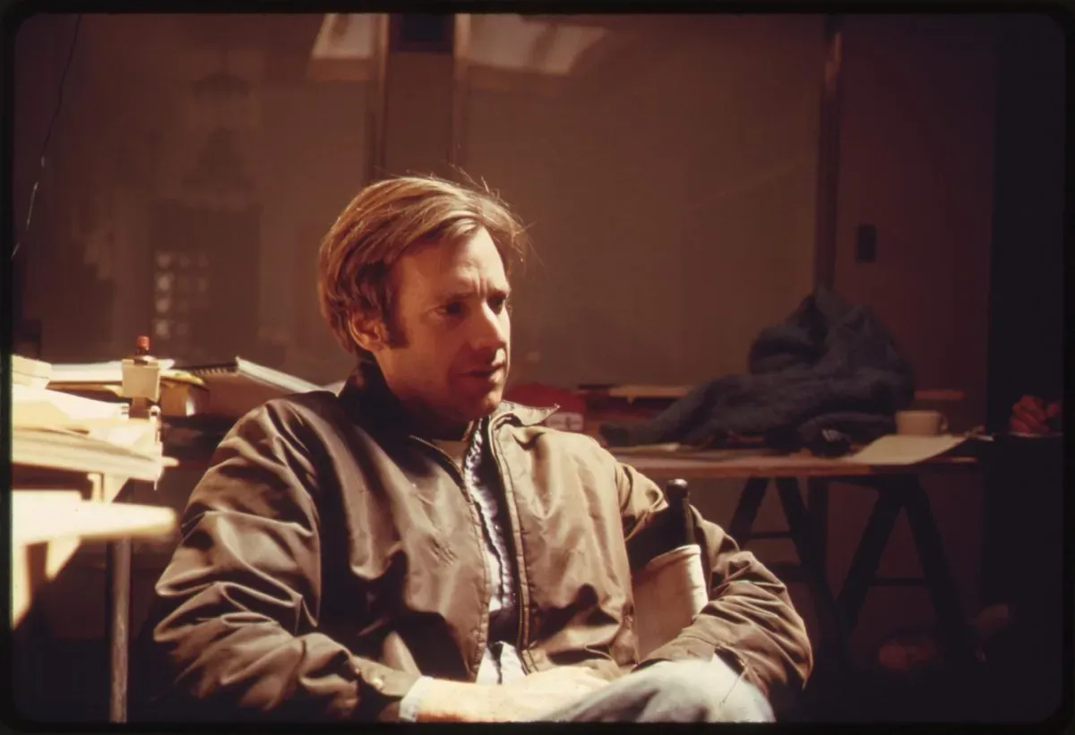 Steve Baer seated in a dim, warmly lit drafting studio, wearing a tan/olive jacket, with drafting tables, papers, a red-capped tube, and a coffee cup visible in the background